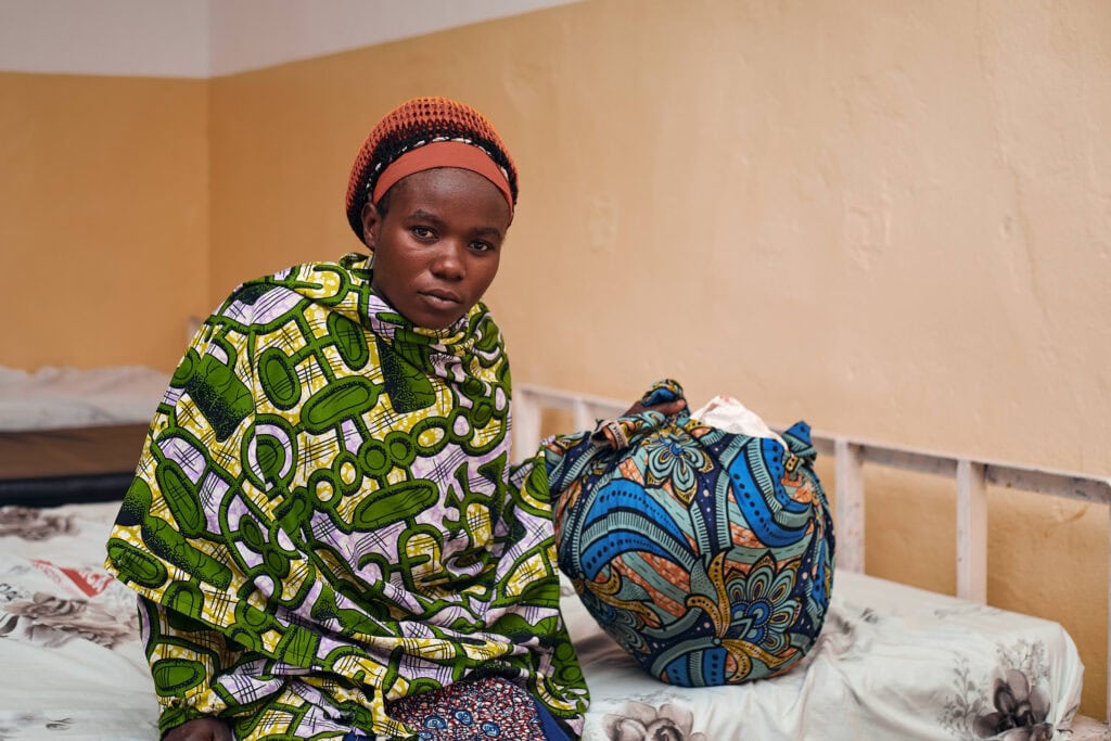 A woman wearing a green and yellow wrapper sits on a hospital bed in the DRC.